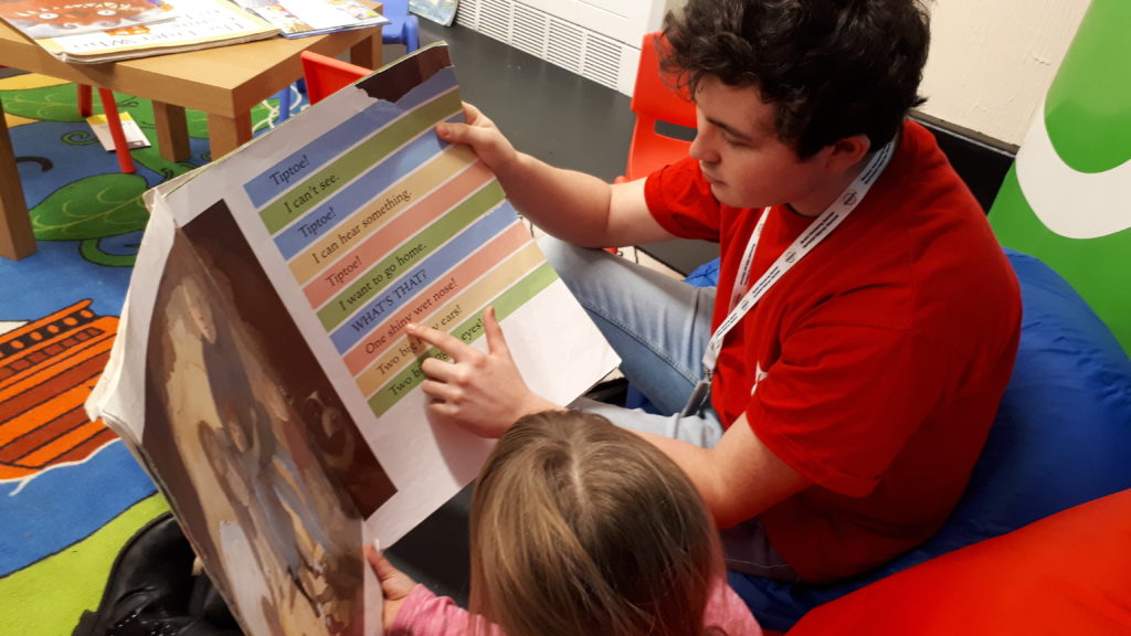 a Swansea university student volunteer in a red tshirt & blue jeans helps a young person learn to read. 